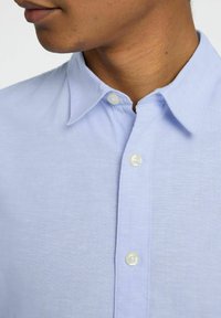 Camicia azzurro chiaro con colletto a punta, realizzata in tessuto tessuto. Presenta bottoni bianchi lungo la patta e una texture sottile.