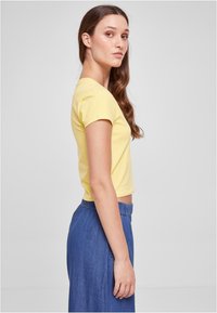 Gul kortermet tettsittende t-skjorte med rund hals, kombinert med et høyt liv blå jeansskjørt, sidevisning. Enkelt design, myk stoff.