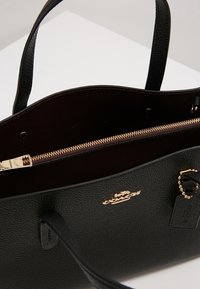 Czarna skórzana torebka o teksturowanej powierzchni, z złotymi akcentami, wewnętrzną kieszenią na zamek i podwójnymi, solidnymi uchwytami. Logo wytłoczone z przodu.