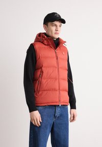 Rød dunvest med hette, med glidelåslommer og glidelås foran, brukt over en svart genser, kombinert med blå jeans.