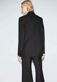 Czarna, dopasowana marynarka o zarysowanej konstrukcji, z bocznymi rozcięciami i trzema guzikami na każdym rękawie, zestawiona z pasującymi spodniami o szerokich nogawkach.