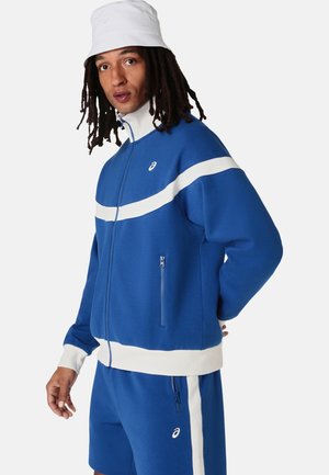 Mand med dreadlocks iført en hvid bucket hat, blå og hvid lynlåstrøje og matchende blå shorts med hvide striber.