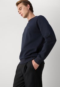 Marinblå sweatshirt i slät tygkvalitet, med rund halsringning och ribbade ärmslut. Inkluderar en liten broderad logotyp på bröstet.