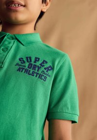 Zelená polokošeľa s krátkymi rukávmi, textúrovaná látka, s modrou vyšitou potlačou „SUPER DRY ATHLETICS“ na prednej strane.