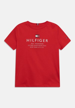 T-shirt rouge à manches courtes avec « HILFIGER » et texte blanc sur la poitrine, petit logo drapeau au-dessus du texte et un autre drapeau sur la manche gauche.