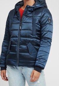Chaqueta acolchada azul marino con capucha, que presenta un cierre de cremallera, puños acanalados y múltiples bolsillos. Textura suave con diseño acolchado.