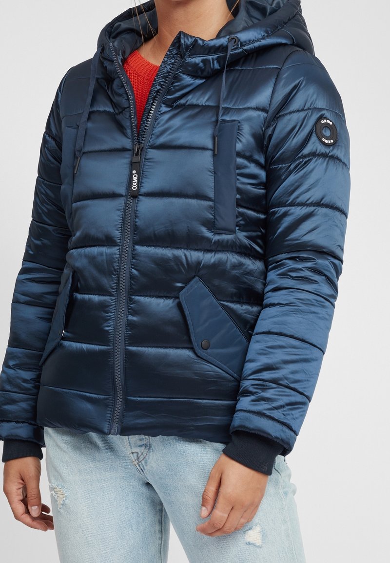 Chaqueta acolchada azul marino con capucha, que presenta un cierre de cremallera, puños acanalados y múltiples bolsillos. Textura suave con diseño acolchado.
