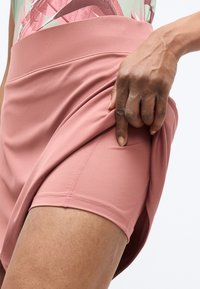 Detailný záber na osobu, ktorá zdvíha ružovú športovú sukňu, aby odhalila pod ňou priliehavé zladené šortky.