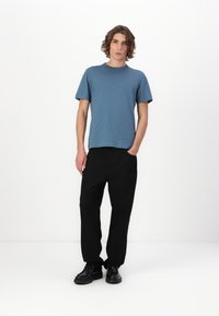 Tricou albastru cu mânecă scurtă, asortat cu pantaloni negri largi; modelul stă cu o mână în buzunar, purtând pantofi negri. Design simplu și curat.