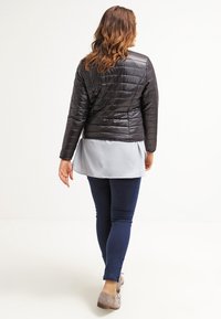 Sort quiltet jakke med en blank overflade, lagd over en lysegrå top, parret med blå jeans og grå fodtøj.