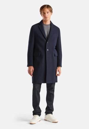 Abrigo de lana navy con solapas anchas, cierre de un solo botón y dos bolsillos frontales, combinado con un suéter gris y jeans oscuros.