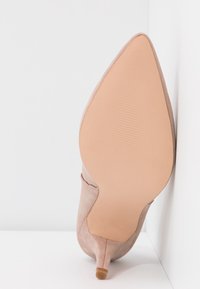 Chaussure beige en daim à bout pointu et petit talon, inclinée verticalement pour montrer la semelle lisse beige clair et la petite pointe du talon sur fond blanc.