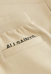 Beżowa bluza z teksturowanej tkaniny, z prążkowanymi mankietami oraz naszywaną kieszenią z napisem "ALLSAINTS" w kolorze czarnym.