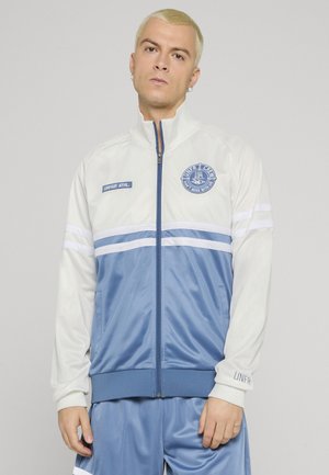 Junger Mann mit kurzen blonden Haaren, der eine weiße und blaue Sportjacke mit Reißverschluss und passende blaue Hose vor einem einfarbigen Hintergrund trägt.