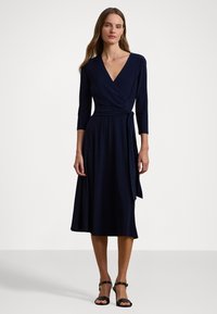 Abito avvolgente blu navy con scollatura a V, maniche a tre quarti e vita legata. Gonna fluida a forma di A che arriva a metà polpaccio. Indossato con tacchi neri.