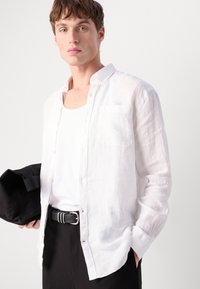 Camisa de linho branca com um bolso, design de botões na frente, mangas longas, usada por cima de uma t-shirt branca, combinada com calças pretas e uma bolsa preta.