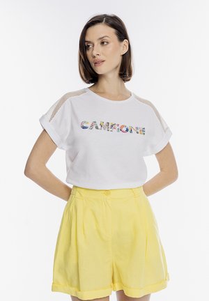 Kobieta o krótkich brązowych włosach ubrana w biały T-shirt z siateczkowymi rękawami i napisem "CAMPIONE", zestawiony z żółtymi szortami z wysokim stanem.