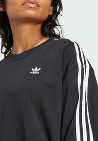 Svart sweatshirt med vitt tre-stripes mönster på ärmarna och liten vit Adidas-logotyp på bröstet, med rund halsringning och mjuk textur.