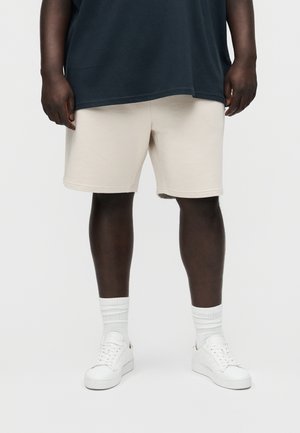 Asmuo dėvintis smėlio spalvos šortus, baltas kojines, baltus sportbačius ir tamsią laisvo kirpimo palaidinę, stovintis priešais paprastą šviesią fono sieną.