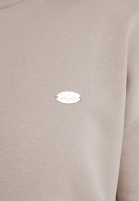 Pull léger beige avec une texture côtelée, un col rond et une étiquette en argent ovale avec du texte gravé sur le devant.