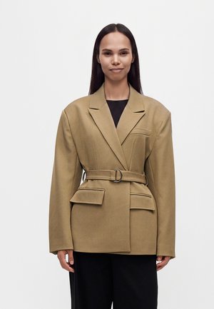 Femme aux longs cheveux foncés portant un blazer ceinturé marron clair oversize et un pantalon noir, debout devant un fond blanc uni.