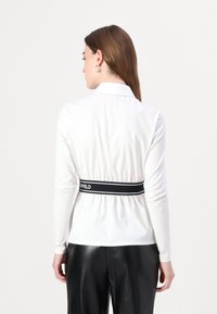 Maglietta bianca a maniche lunghe con vita aderente, caratterizzata da un elastico nero e un accento con logo. Il retro presenta un design con colletto classico.