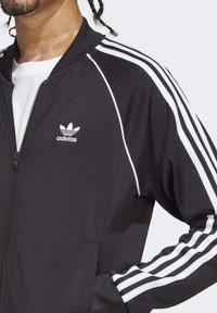 Melna zip-up jaka ar baltām svītrām gar piedurknēm, ar nelielu balto Adidas logo uz krūtīm. Materiāls izskatās gluds.