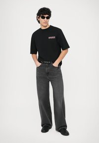 Tricou negru cu logo roșu, asortat cu blugi gri largi și pantofi negri. Păr creț dat impecabil pe spate și ochelari de soare negri.