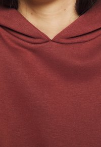 Bordowy kaptur z miękkiej, teksturowanej tkaniny. Posiada zaokrągloną linię dekoltu i klasyczny fason zakładany przez głowę. Brak widocznych logo lub wzorów.