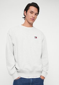 Νεαρός άνδρας που φοράει ανοιχτό γκρι φούτερ με το μικρό λογότυπο της Tommy Jeans στο στήθος και μπλε τζιν, στέκεται μπροστά σε απλή ανοιχτή φόντο.