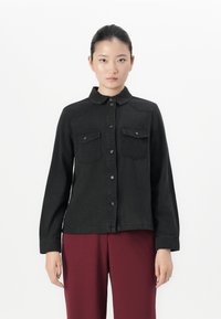 Woman with dark hair pulled back wears a black buttoned shirt jacket with chest pockets and burgundy pants, standing against a white background.