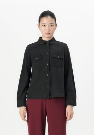 Woman with dark hair pulled back wears a black buttoned shirt jacket with chest pockets and burgundy pants, standing against a white background.
