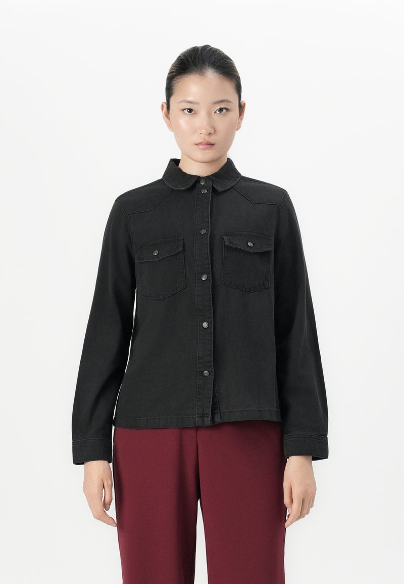 Woman with dark hair pulled back wears a black buttoned shirt jacket with chest pockets and burgundy pants, standing against a white background.