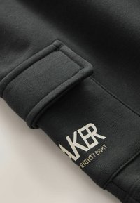 Manche de sweat-shirt noir avec un poignet côtelé et une impression blanche indiquant "AKER EIGHTY EIGHT" près du poignet.