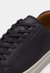 Gros plan sur une chaussure de sport en cuir noir avec des lacets noirs assortis et une semelle blanche, montrant les détails de la couture et la languette rembourrée.