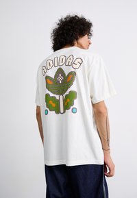 Hvid bomulds-T-shirt med et stort, farverigt pixeliseret grafisk blomstermotiv og "ADIDAS" tekst på ryggen. Korte ærmer, afslappet pasform.