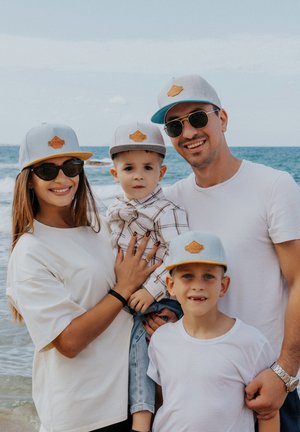 Family portrait at the beach; each wearing light gray caps with a tan patch, sunglasses, and casual white or plaid outfits.