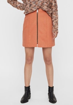 Femme portant un pull marron texturé, une mini-jupe orange avec une fermeture éclair à l'avant, et des bottines noires, sur un fond uni.