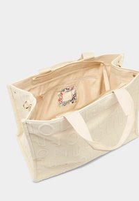 Crèmekleurige boodschappentas van gestructureerde stof met reliëflogo, ritssluiting, binnenzakken en zachte stoffen voering.