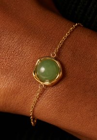 Goldarmband mit einem glatten, runden grünen Stein, der in einer strukturierten, unregelmäßigen Goldfassung eingesetzt ist, verbunden mit einer filigranen Kette.