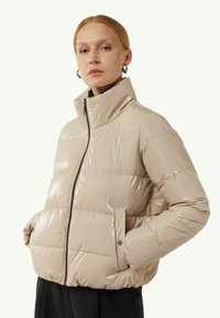 Beżowa kurtka puchowa o błyszczącej powierzchni, wysoki kołnierz, zamek błyskawiczny z przodu i kieszenie boczne; pikowany wzór z wypełnieniem.