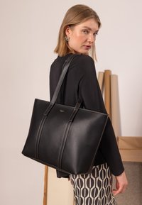 Zwarte leren draagtas met een gestructureerde vorm, voorzien van contrasterende stiksels en twee lange handvatten. Gladde textuur met een subtiel logodetail.