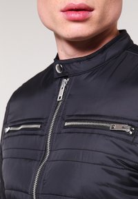 Schwarze gesteppte Jacke mit hohem Kragen, Frontreißverschluss und zwei Brusttaschen mit horizontalen Reißverschlüssen. Glatte Textur.