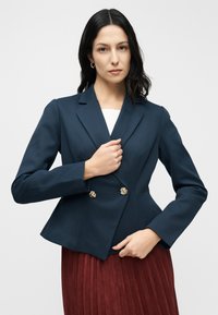 Blazer ajusté bleu marine avec un col cranté, des boutons dorés croisés à double boutonnage, confectionné en tissu texturé. Associé à une jupe plissée bordeaux.