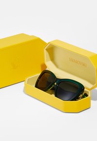 Ochelari de soare Swarovski într-o carcasă dură galbenă, texturată. Ochelarii de soare au rame mari, verzi, cu lentile întunecate. Accente aurii observate.