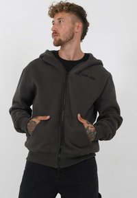 Donkergrijze zip-up hoodie met een capuchon, voorzakken, gestructureerde stof en geborduurd logo op de borst. G gedragen over een zwart shirt.