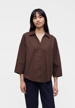 Femme aux longs cheveux noirs portant une chemise marron ample à boutons et un pantalon sombre, debout devant un fond clair uni.
