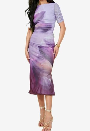 Femme portant une robe midi florale ajustée lavande et violette avec des manches courtes froncées et des sandales à talons hauts dorées avec des lanières.