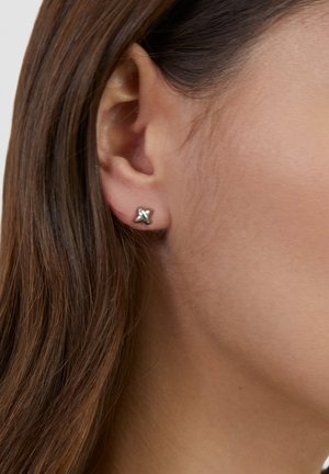 Nahaufnahme eines Ohrs einer Frau mit glattem braunem Haar, die einen kleinen silbernen Ohrstecker in Knotenform am Ohrläppchen trägt.