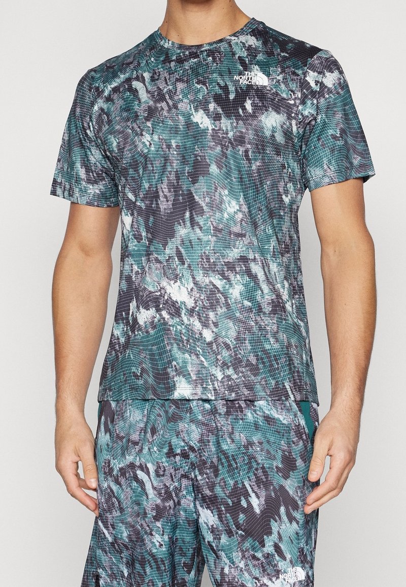 Hombre que lleva una camisa de manga corta y pantalones cortos a juego con patrones abstractos en azul, negro y blanco con ondas y los logos de The North Face.